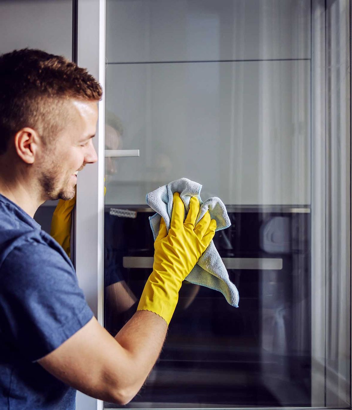 Junger bärtiger, lächelnder, positiver Mann mit Gummihandschuhen, der das Fenster mit einem Tuch putzt. Nach Regen gibt es immer Flecken auf Glas.